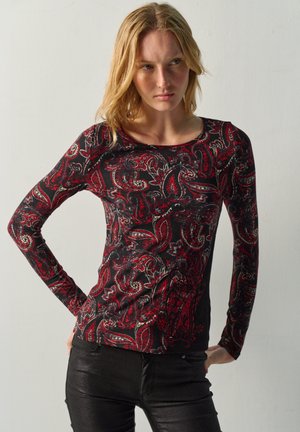 Mujer joven con pelo rubio que lleva una camiseta negra de manga larga con patrones de cachemira rojos y pantalones negros, de pie contra un fondo liso.