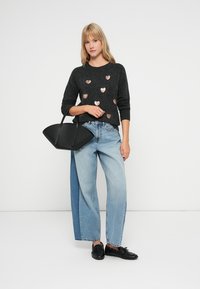 Czarny sweter z metalicznymi zdobieniami w kształcie serca, jasnoniebieskie szerokie dżinsy oraz czarna torebka o strukturalnym kształcie. Czarne buty dopełniają całość.