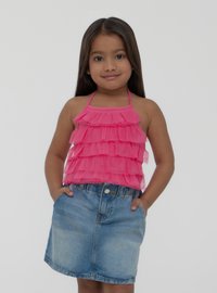 Top halter a balze rosa con volant trasparenti abbinato a una gonna di jeans blu chiaro. La modella ha le mani in tasca e capelli lunghi e scuri.