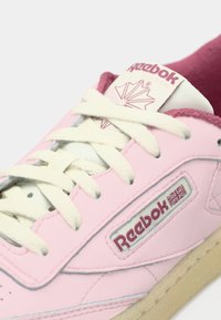 Svijetloružičaste Reebok tenisice s bijelim vezicama, gornji dio od teksturirane kože i gumeni potplat u krem boji. Logo i akcenti u tamnoružičastoj boji.