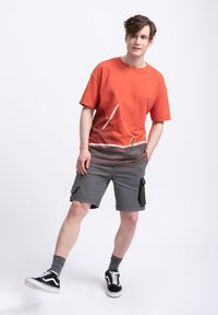 T-shirt en coton rouge avec un design en dégradé et des accents blancs, assortie à un short cargo gris. Le modèle porte des baskets noires et des chaussettes grises.