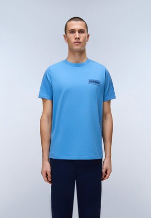 Mężczyzna stoi zwrócony przodem, ubrany w niebieski, krótkim rękawem t-shirt z małym logo na piersi oraz ciemnoniebieskie spodnie na jednolitym tle.