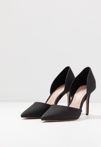 Scarpe nere con tacco alto, punta affusolata, tessuto testurizzato, lati aperti e tacchi a stiletto sottili. Fodera interna di colore chiaro.