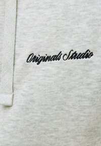 Lys grå sweatshirtstof med en blød tekstur. Har broderet tekst "Originals Studio" i sort. Inkluderer et snoretræk.