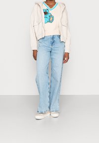 Beige kort jacka över en krämfärgad tröja med en blå björndesign, kombinerad med ljusblå utsvängda jeans och vita sneakers.