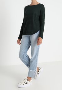 Mörkgrön stickad långärmad topp med texturerade ränder, kombinerad med ljusblå jeans med uppvikta ben och vita sneakers med svarta prickar.