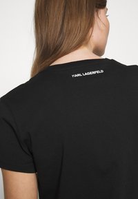 Musta puuvillainen t-paita lyhyillä hihoilla, jossa on pyöreä pääntie ja valkoinen "KARL LAGERFELD" -logo painettuna ylös selässä.
