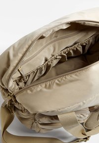 Borsa a spalla in tessuto beige con una finitura testurizzata. Presenta una chiusura con zip, un bordo elastico all'interno e una tracolla regolabile.