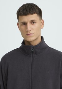 Hall hallitõmblusega jope, millel on kõrge krae, ees tõmblukk ja rinnal tikitud logo. Sile tekstuur ja vaba lõige.