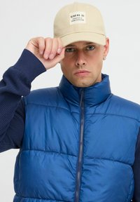 Zila pūkaina veste ar rāvējslēdzēja priekšu, ribotu tumši zilu piedurknēm un augstu apkakli. Beigē beisbola cepure ar logo uz priekšu un plakanu maliņu.