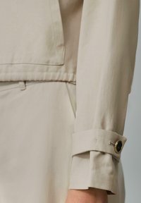 Manche de veste beige sur mesure avec un détail de poignet mettant en valeur un bouton décoratif et des coutures contrastantes, fabriquée en tissu lisse et léger.