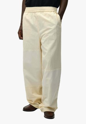 Persona che indossa pantaloni patchwork color crema, larghi, con vita elastica e scarpe di pelle marrone, in piedi con le mani nelle tasche.