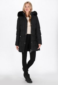 Giacca a vento nera con cappuccio in pelliccia sintetica, dotata di chiusura con zip, tasche laterali e design a diamante trapuntato, abbinata a leggings neri e stivali.