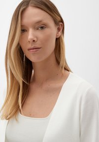Witte trui met een V-hals ontwerp en geribbeld textuur. Model heeft lang, steil haar, zachte gelaatstrekken en een neutrale achtergrond.