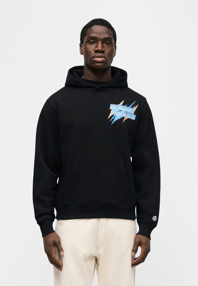 Άνδρας που φοράει μαύρο φούτερ με πολύχρωμο κείμενο "Billionaire Boys Club" στο στήθος και ανοιχτό μπεζ παντελόνι, στέκεται μπροστά σε λευκό απλό φόντο.