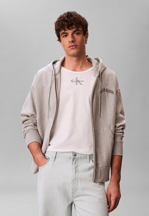 Junger Mann trägt eine hellgraue Calvin Klein Zip-Up-Hoodie, ein weißes Calvin Klein T-Shirt und helle Jeans, steht mit einer Hand in der Tasche.