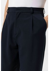 Marineblauwe broek gemaakt van gladde stof met een getailleerd ontwerp, voorzien van voorplooien, zijzakken en een aangesloten taille met ceintuurlussen.