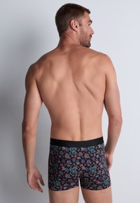 Homme adossé à un mur uni, vu de dos, portant un slip noir avec un motif de récif corallien coloré.