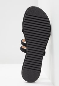 Semelle de sandale noire avec des rainures horizontales pour l'adhérence, montrant une partie de deux lanières noires au-dessus de la semelle.