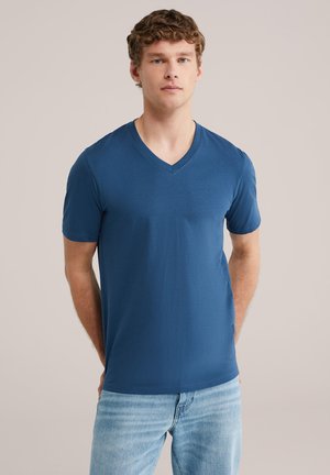 Jonge man met krullend haar, gekleed in een blauw T-shirt met V-hals en lichtblauwe spijkerbroek, staand met zijn handen achter zijn rug tegen een neutrale achtergrond.