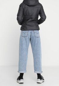 Sort pufferjakke med hætte, parret med lyseblå, opkrøllede denimjeans. Modellen har sorte sokker og sportssko på.