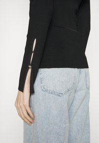 Czarny prążkowany top z długim rękawem z wycięciami na mankietach, ozdobiony srebrnym guzikiem, zestawiony z jasnoniebieskimi dżinsami.