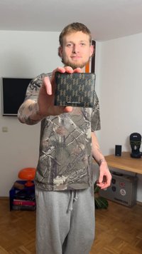 Μαύρο πορτοφόλι με γεωμετρικό σχέδιο σε αποχρώσεις του πράσινου και του καφέ, κρατημένο σε χέρι, εμφανιζόμενο σε ουδέτερο φόντο.