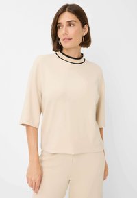 Beige kortærmet top med rund halsudskæring, der har en kontrasterende sort kant. Blødt, glat stof med en afslappet pasform og minimalistisk design.