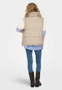 Bēšs pūkains vestes ar augstu apkakli un teksturētiem, vilnas akcentiem, valkāts pār zilpelēku svīainu krekls, kombinēts ar pieguļošām zilām džinsi un brūnām zābakām.