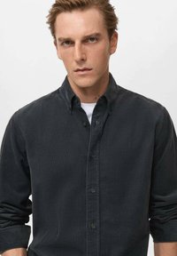 Camisa oscura de botones hecha de una tela suave con un cuello estructurado, que presenta mangas largas y un ajuste holgado. Mangas enrolladas, sin patrones visibles.