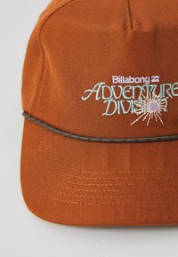 Rozsdabarna sapka fekete díszítéssel a peremén, hímzett "Billabong Adventure Division" felirattal és napfénygrafikával kék és fehér szálakkal.