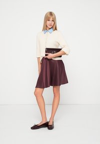 Kremfarget kortermet topp med blå krage, kombinert med en burgunder satin skjørt; modellen holder en mørk clutch med metalliske detaljer.