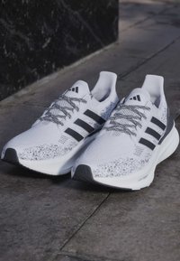 Chaussures de course Adidas blanches et noires avec texture tricotée, bandes noires, lacets à motifs, et semelles intermédiaires "Light Boost" sur pavé en pierre.