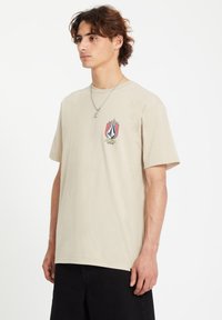 Beige cotton t-shirt with a round neck, short sleeves, and a colorful flame-patterned logo on the left chest. Paired with black shorts.