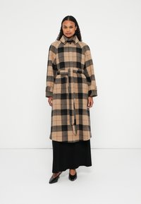 Manteau long à carreaux beige et noir avec une ceinture, un col boutonné et des poches. Porté sur une jupe noire et associé à des talons noirs.