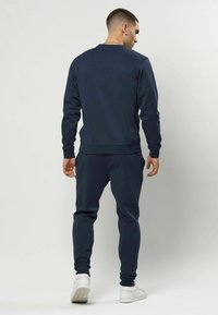 Bărbat purtând un hanorac bleumarin și pantaloni sport, stând cu spatele către înainte, pe un fundal simplu și deschis la culoare.