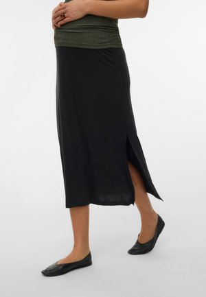 Femme portant une jupe noire mi-longue fendue sur le côté, associée à des ballerines noires et un haut côtelé vert, les mains reposant sur sa taille.