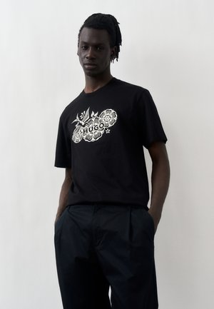 Joven con rastas que lleva una camiseta negra con diseño floral blanco y texto "HUGO", de pie con las manos en los bolsillos frente a un fondo liso.