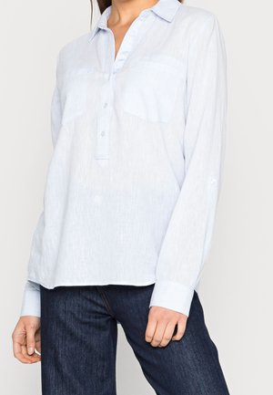 Kobieta ubrana w jasnoniebieską koszulę z długim rękawem, kołnierzykiem i guzikami oraz ciemnoniebieskie dżinsy, stojąca na jednolitym tle.