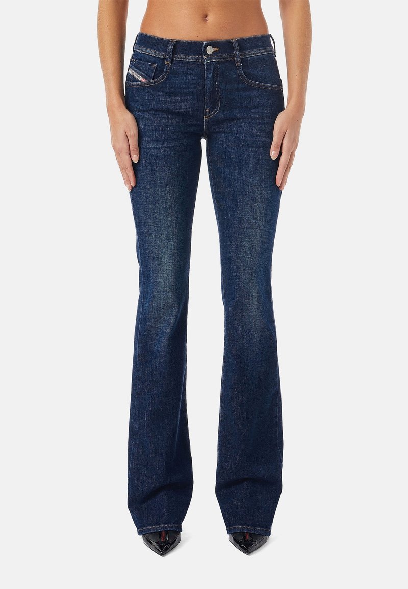 Femme portant un jean bootcut bleu foncé avec poches avant et fermeture par boutons, debout avec les mains détendues le long du corps.