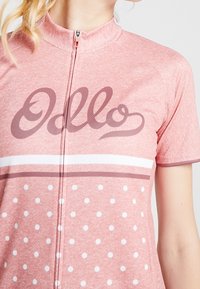 Różowa koszulka kolarska z krótkim rękawem, z zamkiem z przodu, białe kropki na dolnej części oraz grafika "Ollo" w kolorze bordowym na klatce piersiowej.