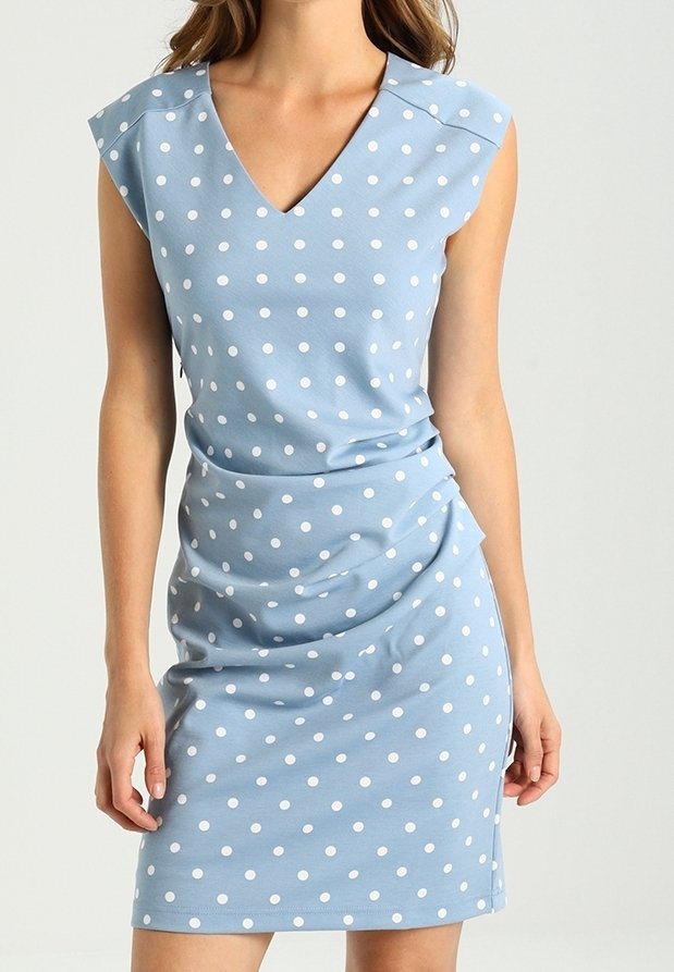 Femme portant une robe sans manches bleu clair à pois blancs avec un décolleté en V, posant devant un fond uni.