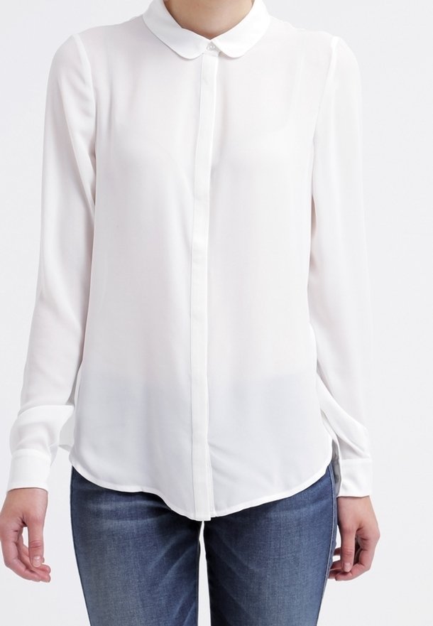 Blusa blanca de manga larga con un frente abotonado, cuello redondeado y tejido suave. Presenta un dobladillo curvado y un ligero brillo. Se lleva con vaqueros azules.