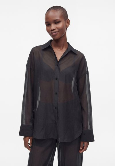 Personne portant une chemise noire transparente à boutons et un pantalon transparent assorti, debout devant un fond blanc, souriant légèrement.