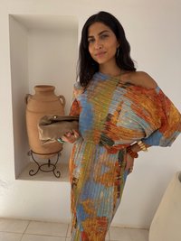 Színes, plisszírozott ruha kék, narancs és zöld árnyalatokban. Kicsi barna clutch táskával a kezében. Egyszerű fülbevalók, gyöngyből készült karkötő a csuklóján.