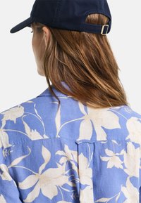 Blå skjorte med blomstermønster og kremfargede blomster, med knapp-ned krage og detalj med bakfold. Laget av bomull, med en uformell design. Mørkeblå caps på hodet.