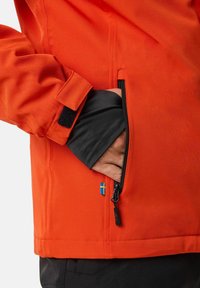 Giacca arancione con tasca con cerniera, fodera interna nera, polsini in velcro e un piccolo dettaglio con bandiera blu e gialla sul lato.