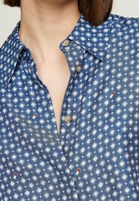 Nainen, joka on pukeutunut siniseen painettuun kauluspaitaan, jossa on geometrinen timanttimuotoilu, ja joka on osittain avattu ylhäältä.