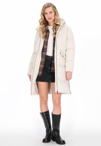 Vrouw in een beige pufferjack over een geruit overhemd, witte top, zwarte rok en zwarte kniehoge laarzen, staande tegen een witte achtergrond.