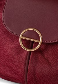 Bordeauxfarbene synthetische Handtasche aus strukturiertem Material, mit einem runden goldenen Hardware-Akzent und einem Klappenverschluss-Design.